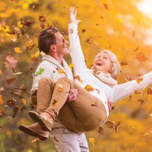 older couple enjoying the fall season. Husband carrying wife through falling leaves.